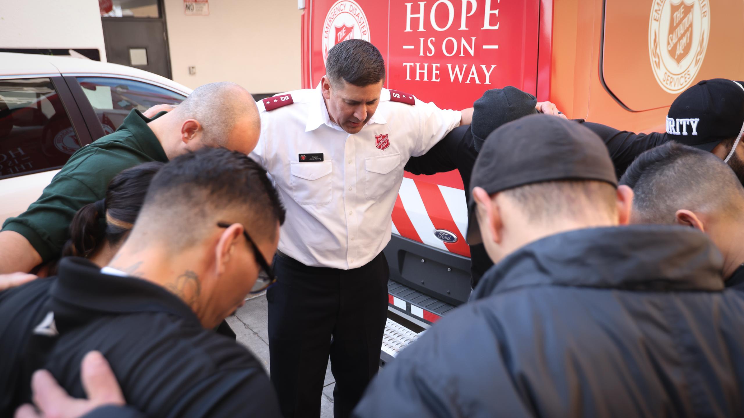 Eight people including a Salvation Army officer stand in a circle, hands around each others' shoulders and heads bowed in prayer. In the background a Salvation Army truck shows the message 'Hope is on the way'.